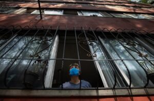 Una mujer con mascarilla mira por la ventana durante el confinamiento domiciliario provocado por la pandemia global de Coronavirus