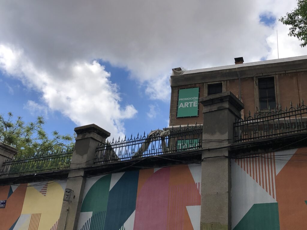 Exterior del edificio de La Tabacalera, en Madrid. Es de día, detrás de un muro con pintadas de colores, un edificio de apariencia abandonada con un cartel en una de sus ventanas: 'Promoción del arte'.