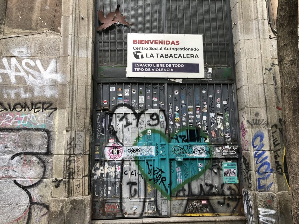 Entrada del CSA La Tabacalera. Es de día, la puerta esta grafiteada, con pegatinas y mensajes reivindicativos. Un cartel de fondo blanco encima de la puerta: 'Espacio libre de todo tipo de violencia'.