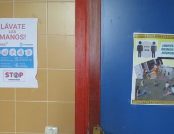 2 carteles, uno en un apuerta azul y otra en una pared de azulejo naranja que muestran medidas para no contagiar