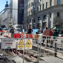 Calle Gran Vía a la altura de la calle Montera. Se ven las vallas de las obras y personas caminando entre ellas. De día.