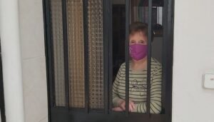 Una señora mayor asomada con mascarilla asomada a la puerta de su casa.