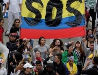Plano picado y frontal de un grupo de unas veinte personas colombianas manifestándose con una gran bandera de Colombia en la que aparece escrito en letras mayúsculas SOS.