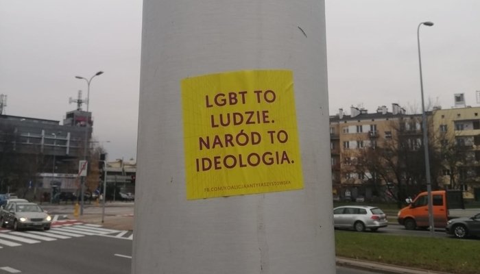 Pegatina amarilla en un poste con texto en polaco que dice: "Los LGTBI+ son personas. La nación es ideología". Ciudad de fondo.